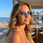 Mulher com os óculos Mionelle Facet Sky Blue ao ar livre, com praia e palmeiras ao fundo