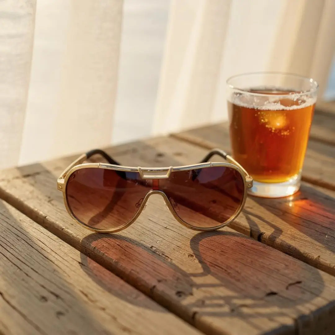 Óculos de Sol Aviador Vintage Masculino e Feminino Mionelle Tesla Marrom sobre uma mesa de madeira ao lado de um copo com bebida